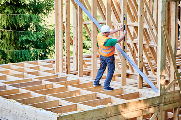 Carpenter constructing two-storey wooden frame house. Back view of man inspects walls for levelness using spirit level, wearing protective overalls, helmet and vest. Concept of modern construction.