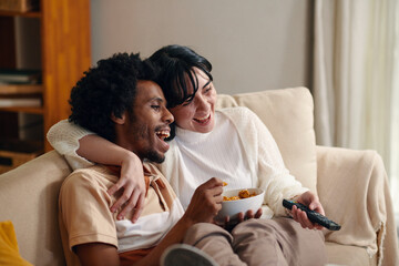 Young laughing man with bowl of popcorn and his wife with remote control sitting on couch in front...
