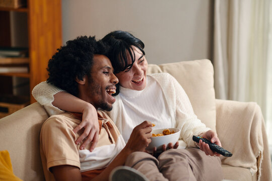 Young laughing man with bowl of popcorn and his wife with remote control sitting on couch in front of TV set and watching sitcom