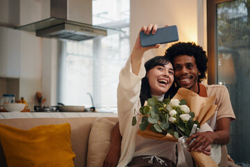 Happy wife with bouquet of flowers holding smartphone in front of herself and taking selfie with...