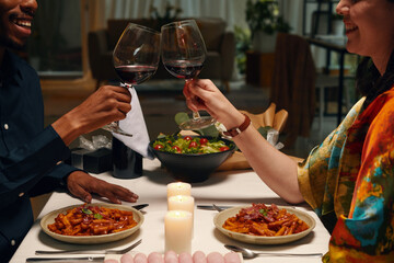 Cropped shot of happy young intercultural valentines clinking with glasses of red wine while sitting by served table with homemade food