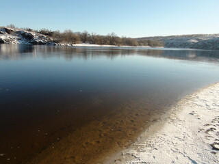 A panorama of the transparent water surface of the wide Dnieper River, through which a clean sandy bottom surrounded by a snow-covered shore shines through.