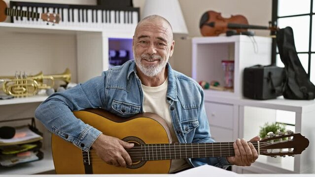 A senior man with a beard in a casual office plays guitar while gesturing to stop with his hand.
