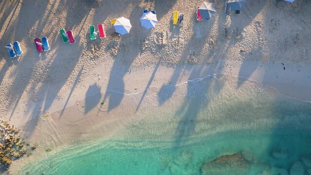 Crystal clear ocean waves crash gently on golden sand beach with bright colored chairs and umbrellas, aerial top down