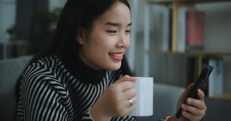 Portrait of Happy young asian woman sit on sofa enjoy chatting on mobile phone while drinking...