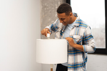 Smiling young African American man, master replacing light bulb in floor lamp, standing in room at...