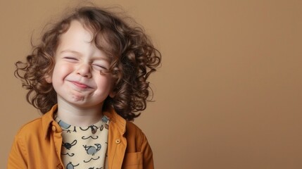 A young child with curly hair wearing an orange shirt with a playful design smiling brightly with eyes closed against a neutral beige background.