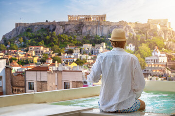 A tourist man sitting by the pool enjoys the view over the old town of Athens, Greece, and the Acropolis during his summer vacations