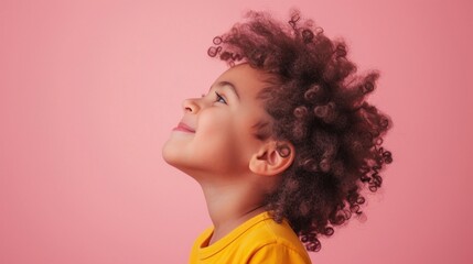 A young child with curly hair wearing a yellow shirt looking up with a smile against a pink background.