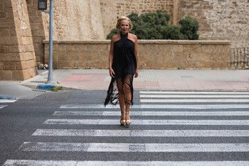 Mature, blonde, beautiful woman crosses the pedestrian crossing. The woman is wearing a black dress...