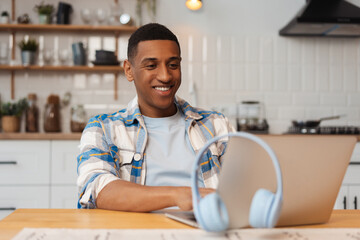 Portrait of smiling positive African American man sitting at workplace, using laptop, ordering
