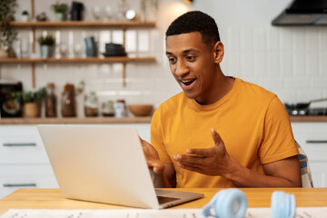 Portrait of confident young African American man sitting in modern kitchen at home, using laptop,...