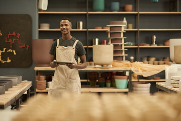 Young African potter with a tablet smiling in a large ceramics workshop - obrazy, fototapety, plakaty