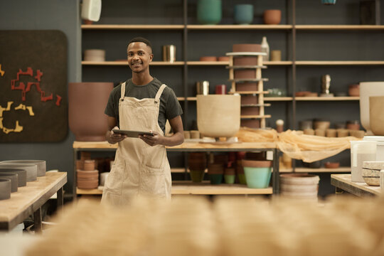 Young African potter with a tablet smiling in a large ceramics workshop
