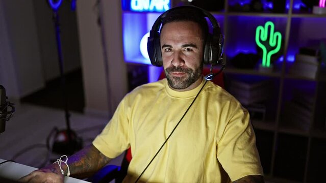 Bearded man wearing headphones in a neon-lit gaming room looking at screen