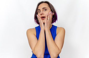 Fototapeta premium Surprised woman looks with open mouth. Hands near cheeks. Blue dress. White background, isolated.