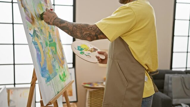A tattooed artist in an apron stands painting on an easel in a bright, window-lit studio.