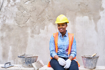 Portrait of smiling young female bricklayer amidst buckets sitting against wall at construction site