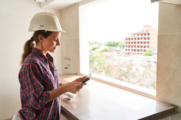 Side view of young female contractor wearing hardhat using tablet PC in incomplete house