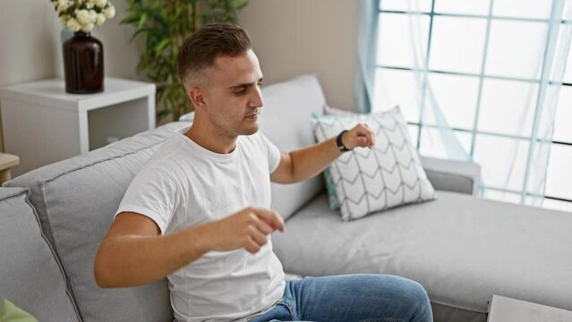 A young hispanic man in casual attire relaxes on a modern sofa in a well-lit living room, portraying a sense of calm domesticity.