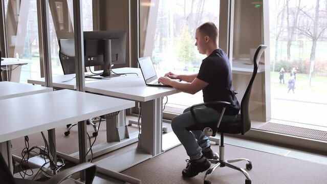 Business concept. Young european man is working in the office on a laptop. Coworking center for entrepreneurs. An IT specialist writes code. SEO manager. Open space with big floor-to-ceiling windows