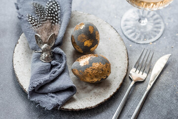 able setting. A fashionable concrete plate with a rabbit on a napkin, Easter eggs with gilding, feathers on a gray background. The concept of a Happy Easter holiday for cafes and restaurants.