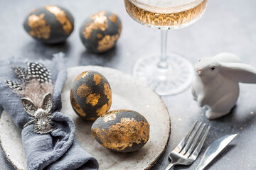 Fototapeta na wymiar able setting. A fashionable concrete plate with a rabbit on a napkin, Easter eggs with gilding, feathers on a gray background. The concept of a Happy Easter holiday for cafes and restaurants.