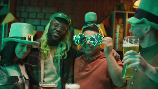Medium footage of cheerful multiracial friends wearing green hats dancing near table in tavern and clinking glasses of emerald lager while celebrating Saint Patricks Day - Powered by Adobe