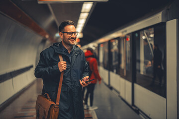 A happy mid adult employee using his phone, ready to get on the train to arrive to his work destination - obrazy, fototapety, plakaty