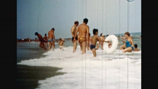 People crowd relax on sea beach in sunny summer. Parents, children with inflatable rings enjoy vacation in ocean shore. Many people in water. City public beach. 1980s archive. Old film. Archival video