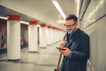 A happy adult businessman leaning on a wall and waiting for a train on a subway station while using...