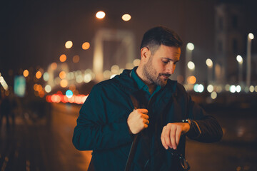 A tired mid adult male employee checking the time on his wristwatch and walking back home during night