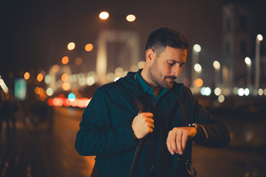 A tired mid adult male employee checking the time on his wristwatch and walking back home during night - Powered by Adobe