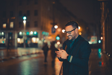 A cheerful adult businessman with glasses walking home from work in the evening and typing text...