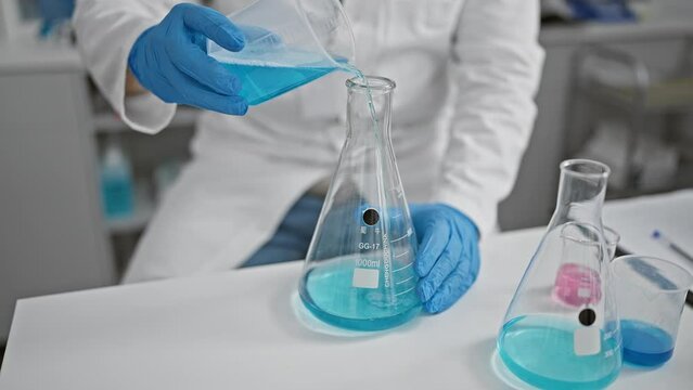 A scientist in a lab coat pouring a chemical from a beaker into a flask indoors.