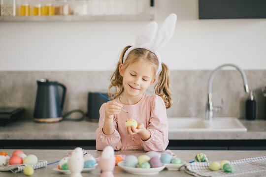 Happy cute little girl decorating Easter eggs using brush and paint on the kitchen.