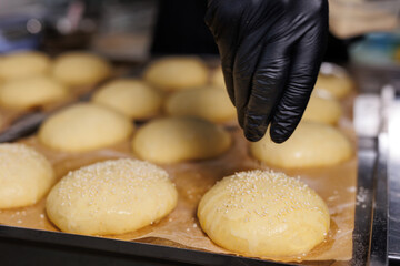 Close-up of the chef's gloved hands, who sprinkles sesame seeds on burger buns before baking.