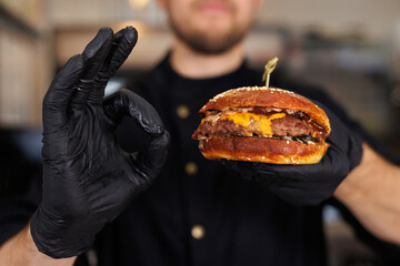 Cook in black gloves holds a burger in his hands and shows an ok sign.