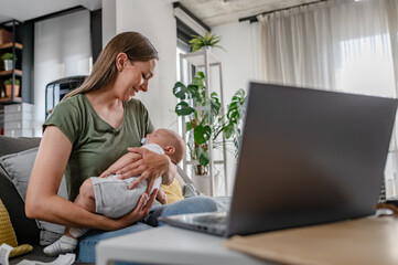 Young new mom holding cute baby in her arms at home while working on a computer