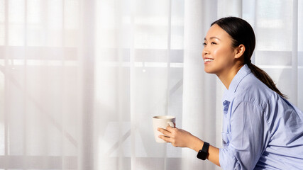 Cheerful asian lady drinking coffee, sitting next to window