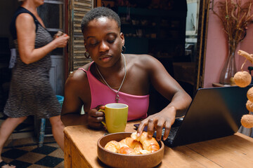 young afro woman at home studying with laptop taking croissant from bowl on table