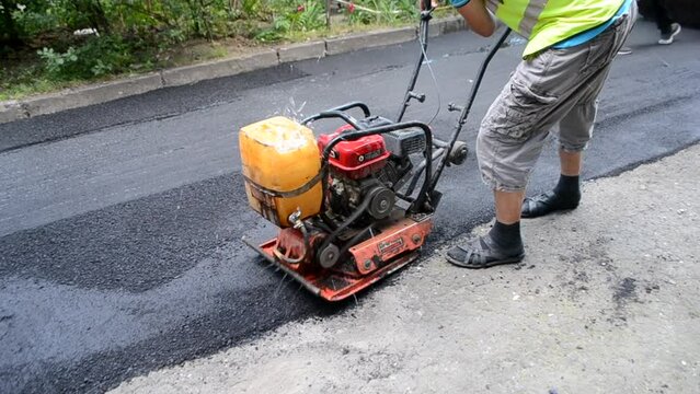 Worker man manually compacts an asphalt concrete mixture with a old vibrating plate while paving road. Compacting tamping asphalt concrete with a manual hand rammer. Asphalting Street paving equipment