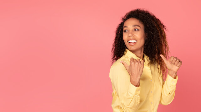 Young african american woman points back to free space on pink backdrop