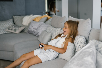 Child girl plays a video game console using a joystick or controller, sitting at home on the couch,...
