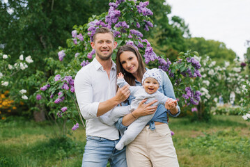 Obraz premium Mother, father and little son are walking in a spring blooming park. A happy mom and dad with a small child are having fun near the lilac. The concept of a happy family.