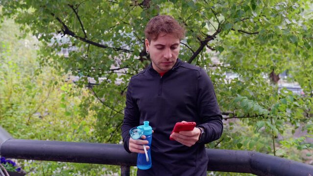 An athletic Caucasian man tired of exercising takes a break to drink water while using a cell phone app. outdoors.