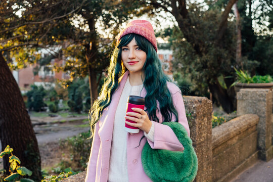 Stylish young smiling hipster woman with color hair walking on street in pink outfit with reusable coffee cup wearing coat, knitted hat, fur bag, happy mood, seasonal fashion, Barbiecore style trend