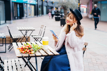 Stylish young smiling woman enjoying coffee from a reusable cup, listening to music, podcast using wireless earbuds and phone in street cafe. Fresh tulips bouquet on table. Springtime street fashion.