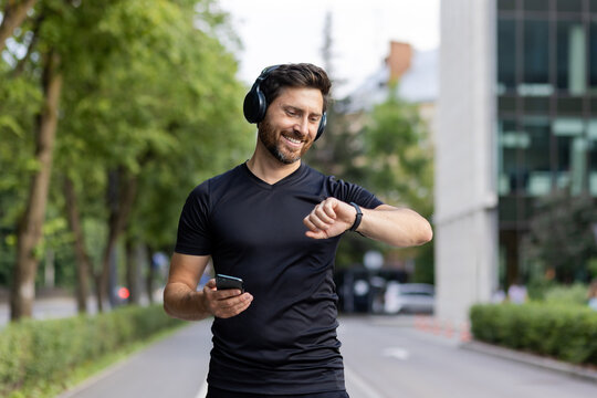 Smiling young man doing sports and running. Standing outside in the city wearing headphones, holding a mobile phone and looking at a smart watch
