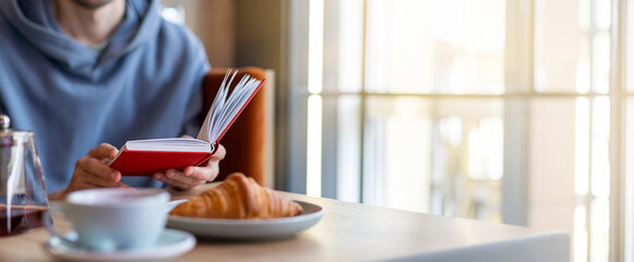 Man reading a book in a cafe with a cup of coffee and a croissant on the table